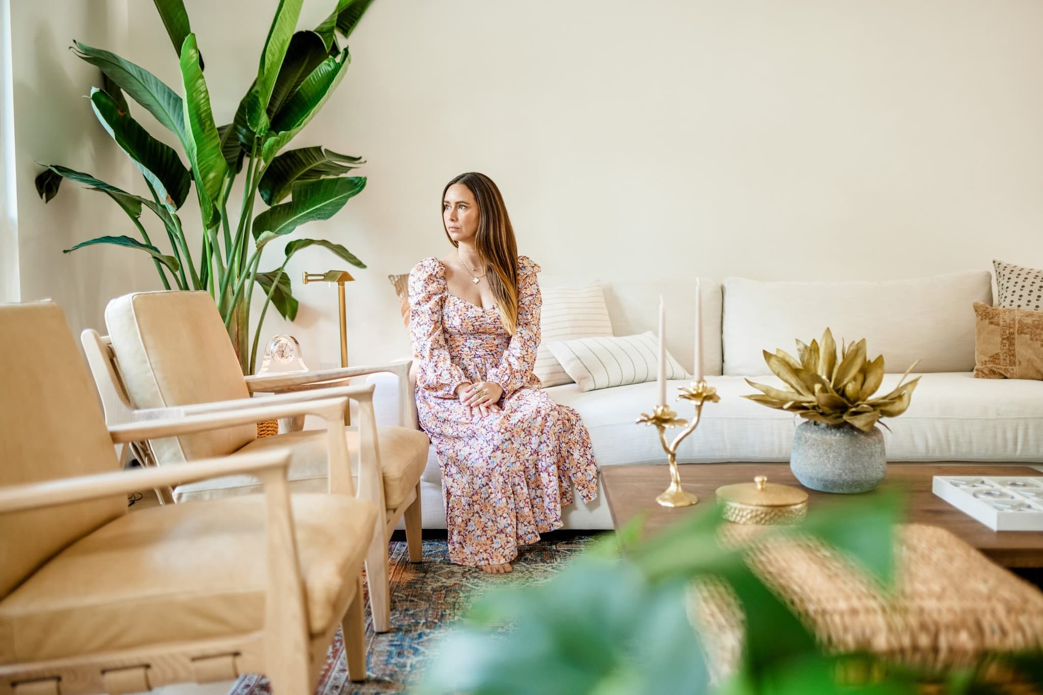 Paula sitting on a white sofa in a bright living room with wood chairs and a tall plant