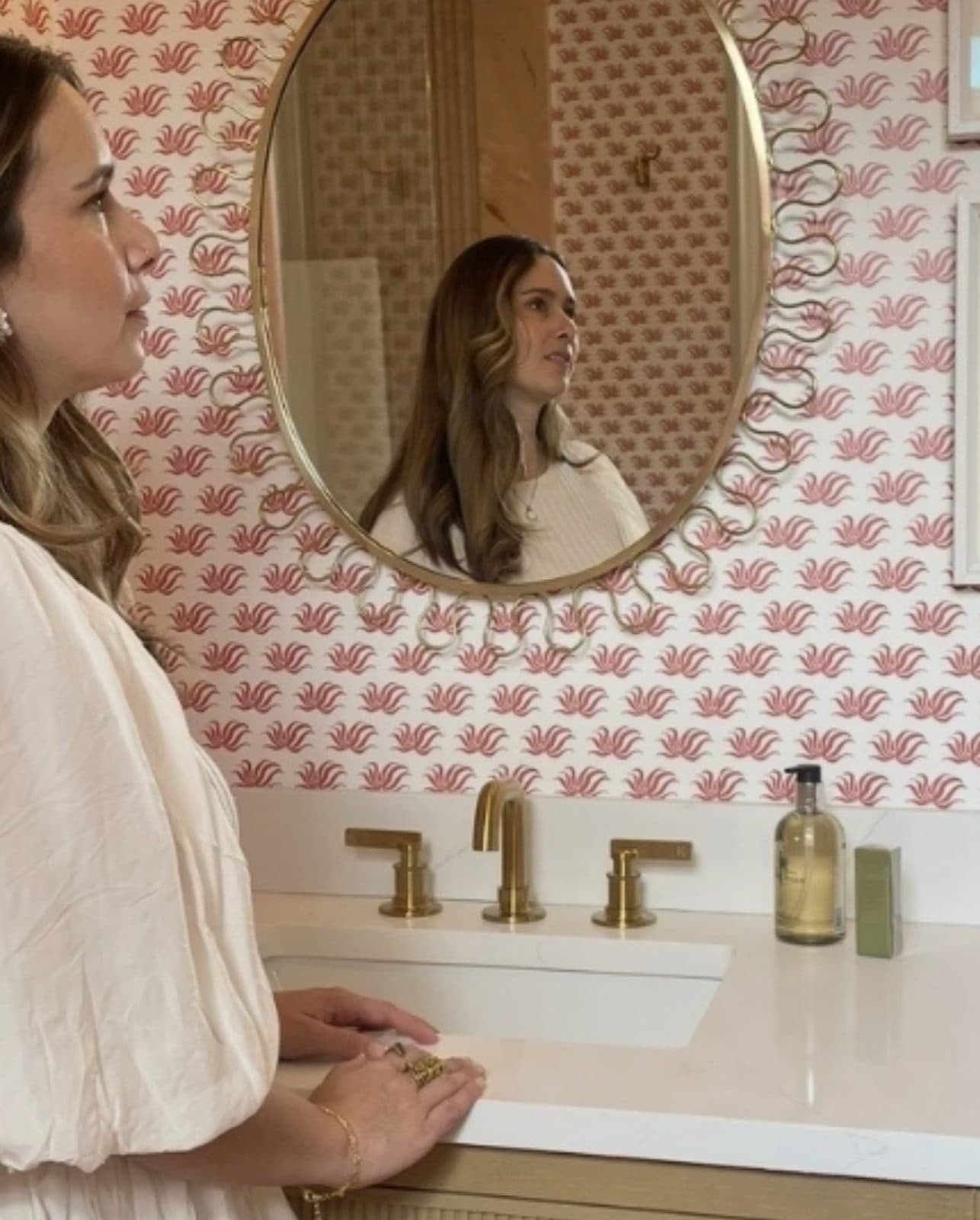 Paula looking into a round gold mirror in a bathroom with pink patterned wallpaper